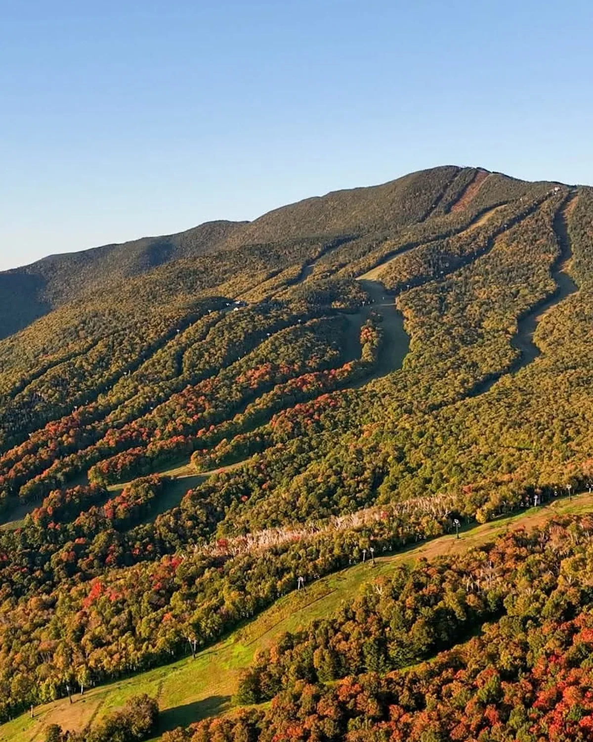 Ski trails at Sugarbush cutting down a green mountainside in early fall, with red maples along the edges under a clear sky